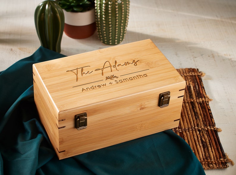 May include: A rectangular bamboo box with a hinged lid and metal clasps. The lid is engraved with the words "The Adams" above "Andrew + Samantha". The box is light brown and sits on a dark teal fabric.