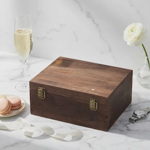 May include: A wooden box with a hinged lid and metal clasps. The box is engraved with a name and date. A glass of champagne, macarons, a watch, and a white flower are also in the image.