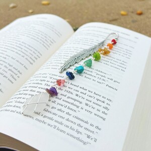 May include: A silver feather bookmark with rainbow colored stones and a white seashell. The bookmark is resting on an open book with text about gossiping and criminals. The book is on a sandy beach.