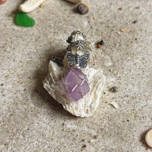 以下が含まれることがあります： ラベンダー色のシーグラスの石が付いたシルバーの蝶のペンダントネックレス。白いサンゴの上に置かれています。ネックレスは、貝殻とヒトデのある砂の表面にあります。