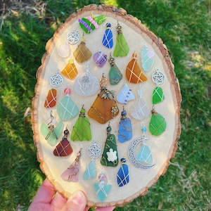May include: A collection of handmade pendants and charms, featuring sea glass and wire wrapping. The pendants come in various colors, including green, blue, orange, and purple. Some charms have star, moon, and other decorative elements. The items are displayed on a wooden slice.