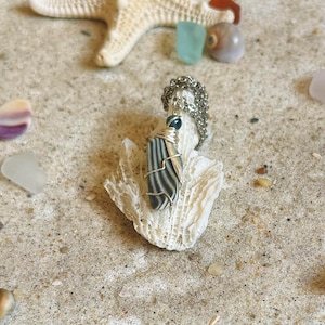 May include: A handcrafted pendant necklace featuring a striped stone wrapped in silver wire. The pendant hangs from a silver chain and rests on a piece of white coral. The background includes a starfish, seashells, and sea glass.