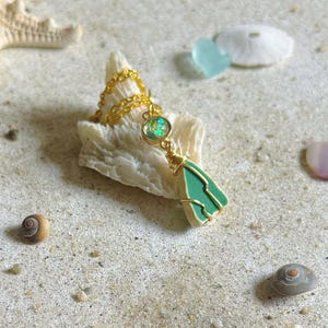 May include: A gold chain necklace with a pendant featuring a green stone wrapped in gold wire. The necklace rests on a white seashell, surrounded by sand, seashells, and a starfish. The pendant has a round, iridescent stone.