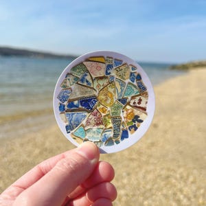 May include: A round, white sticker with a colorful mosaic design made from broken pieces of china. The mosaic is made up of blue, white, yellow, green, and pink pieces. The sticker is held in a hand against a blurred background of a beach.