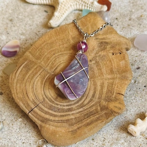 May include: A handcrafted necklace featuring a polished, purple stone pendant wrapped in silver wire. The pendant hangs from a silver chain and is accented with a small, round, purple bead. The necklace rests on a wooden slice, with a starfish and seashells in the background.
