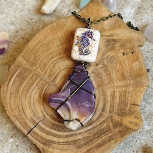 May include: A handcrafted necklace featuring a seahorse pendant and a purple stone. The seahorse pendant is rectangular with a seahorse illustration. The purple stone is wire-wrapped. The necklace is displayed on a wooden slice with a starfish in the background.