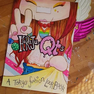 May include: A book cover with a cartoon illustration of a girl with red hair wearing a rainbow shirt and blue jeans. The title of the book is "Harajuku QTs" and the subtitle is "A Tokyo fashion lookbook".