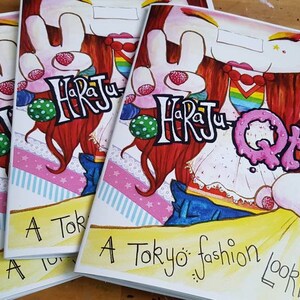 May include: A stack of four colorful paperback books with the title "Harajuku QTs: A Tokyo fashion look book" on the front cover. The cover features a cartoon illustration of a girl with red hair wearing a white dress and blue jeans.