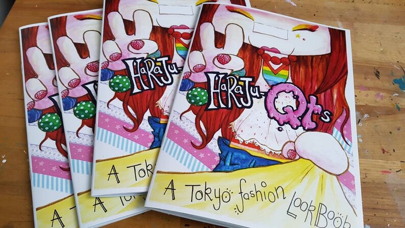 May include: A stack of four colorful paperback books with the title "Harajuku QTs: A Tokyo fashion look book" on the front cover. The cover features a cartoon illustration of a girl with red hair wearing a white dress and blue jeans.