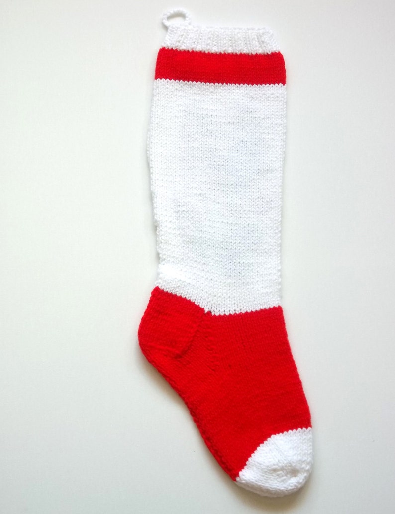 May include: A white knitted stocking with a red band at the top and a red toe. The stocking has a loop at the top for hanging.