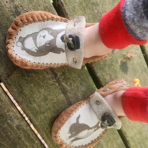 May include: A pair of brown suede moccasin-style slippers featuring a white top panel with a black and white cat illustration. Each slipper has a light beige strap across the instep secured with a black plastic toggle and a cord drawstring.