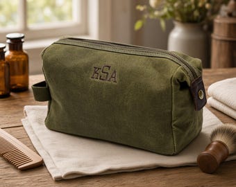 May include: An olive green canvas wash bag with a zipped top closure and a side handle. The front of the bag features the embroidered letters KSA in a dark thread. The bag has a rectangular shape with a brown leather accent on the side.