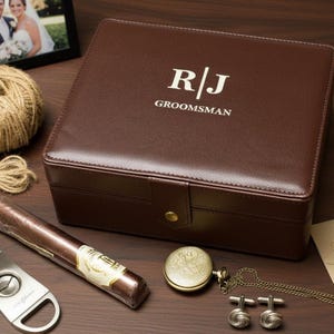 Op de afbeelding: Een bruine leren doos met de initialen "R/J" en het woord "Groomsman" in het wit. De doos is omgeven door een sigaar, een horloge, manchetknopen, een pen, een "Thank You"-kaart en een fotolijst. Een bol touw en een strik zijn ook aanwezig.