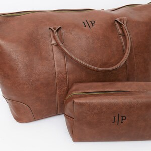 May include: A brown leather travel bag set with a large duffel bag and a smaller toiletry bag. Both bags have a zipper closure and a top handle. The bags are personalized with the initials "J|P" in black.