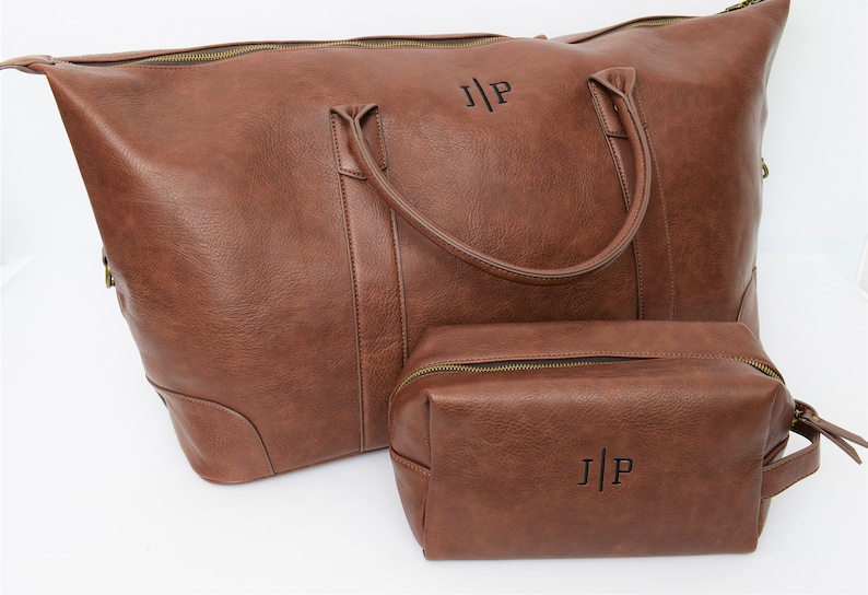 May include: A brown leather travel bag set with a large duffel bag and a smaller toiletry bag. Both bags have a zipper closure and a top handle. The bags are personalized with the initials "J|P" in black.