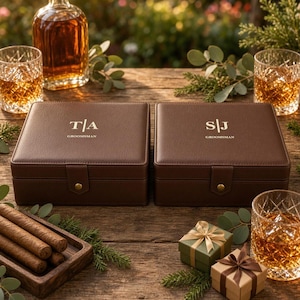 Pode incluir: Duas caixas quadradas de couro castanho com fechos de pressão dourados. Uma caixa está gravada com T|A e GROOMSMAN, a outra com S|J e GROOMSMAN. As caixas estão rodeadas por uma garrafa de vidro com líquido âmbar, três copos de cristal, charutos e duas pequenas caixas de presente.