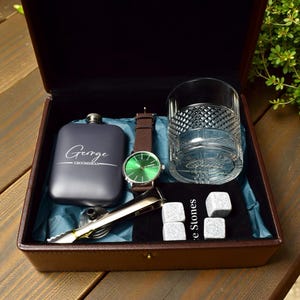 May include: A dark brown leather gift box containing a flask with "George GROOMSMAN" text, a watch with a green face and brown strap, a rocks glass, ice stones, and tongs. The box is lined with blue fabric.