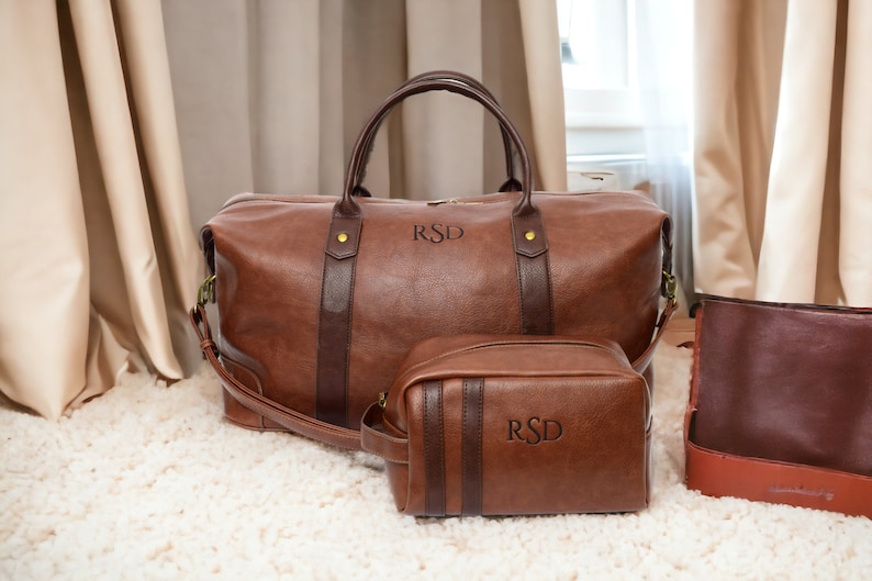 May include: Two brown leather travel bags with the initials "RSD" embossed on them. The larger bag has a top handle and a shoulder strap. The smaller bag is a toiletry bag.