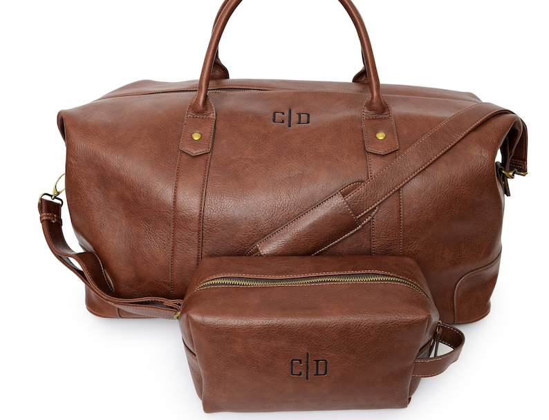 May include: A brown leather travel bag with a matching brown leather toiletry bag. Both bags have a zipper closure and a monogrammed "C|D" on the front.