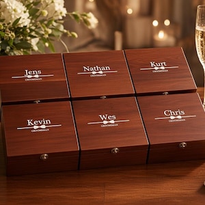 Pode incluir: Seis caixas retangulares de madeira escura com tampas articuladas, cada uma com um fecho prateado e texto gravado em branco incluindo nomes como Jens, Nathan, Kurt, Kevin, Wes e Chris, a palavra GROOMSMAN e um ícone de gravata borboleta.
