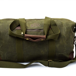 May include: A green canvas duffel bag with brown leather accents and a personalized monogram that reads "KSA". The bag has a top zipper closure, two top handles, and an adjustable shoulder strap.