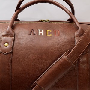 May include: A brown leather duffel bag with a shoulder strap and a zipper closure. The bag has the letters "ABCD" embroidered on the front in different colors.