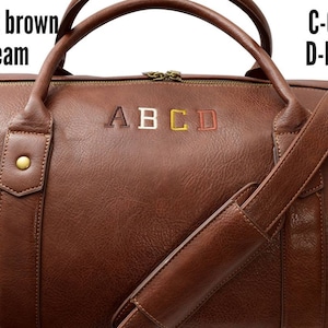 Pu&ograve; includere: Una borsa da viaggio in pelle marrone con una tracolla. La borsa ha una chiusura a zip ed &egrave; monogrammata con le lettere A, B, C e D in diversi colori. Le lettere sono ricamate in marrone scuro, crema, oro e marrone.