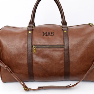 Pu&ograve; includere: Borsa da viaggio in pelle marrone con manico superiore e tracolla. La borsa ha una tasca con cerniera sul davanti e le iniziali "MAS" sono ricamate sul davanti.