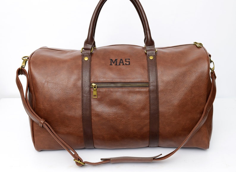 Pu&ograve; includere: Borsa da viaggio in pelle marrone con manico superiore e tracolla. La borsa ha una tasca con cerniera sul davanti e le iniziali "MAS" sono ricamate sul davanti.