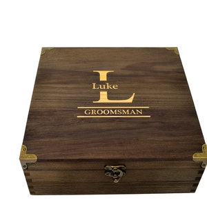 Può includere: Una scatola di legno marrone scuro con angoli in ottone e un chiavistello in ottone. La scatola è incisa con la lettera "L" e le parole "Luke" e "Groomsman".