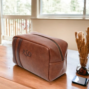 May include: A brown leather travel toiletry bag with a zipper closure and a black leather strap. The bag is monogrammed with the initials "RSD".