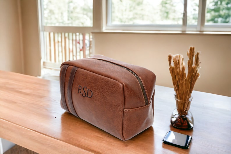 May include: A brown leather travel toiletry bag with a zipper closure and a black leather strap. The bag is monogrammed with the initials "RSD".