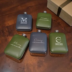May include: Five personalised hip flasks in shades of green and grey, each with a silver cap. The flasks are engraved with names and titles like "Best Man" and "Groomman". A gift box with a ribbon is in the background.