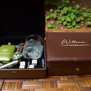 May include: A brown leather gift set containing a watch, hip flask, rocks glass, pen, and whiskey stones. The flask is olive green with initials and the word "Groomsman". The watch has a brown leather strap.
