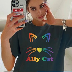 Puede incluir: Camiseta negra con un gráfico de gato arcoíris y el texto "Ally Cat". El texto "Size Up For Trendy Oversized Look" está en la parte inferior de la imagen.