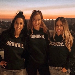 May include: Three friends wearing black hoodies with the words "Ginger", "Brownie", and "Blondie" printed on them. They are standing in front of a sunset.