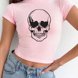 May include: A pink cropped t-shirt with a black skull graphic wearing sunglasses. The skull has a toothy grin.