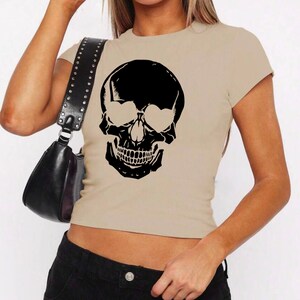 May include: A beige cropped t-shirt with a black skull graphic wearing sunglasses.