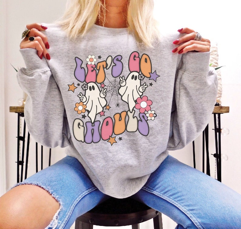 May include: A light grey crew neck sweatshirt featuring the phrase "Let's Go Ghouls" in a retro font. The design includes two cartoon ghosts, flowers, stars, and a spiderweb. The sweatshirt is a casual style.