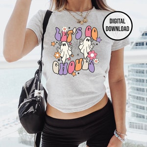 May include: A light grey t-shirt with the text "Let's Go Ghouls" in a retro font, featuring two cartoon ghosts, flowers, and stars. A black circle with "DIGITAL DOWNLOAD" is in the upper right corner. The shirt is part of a Halloween theme.