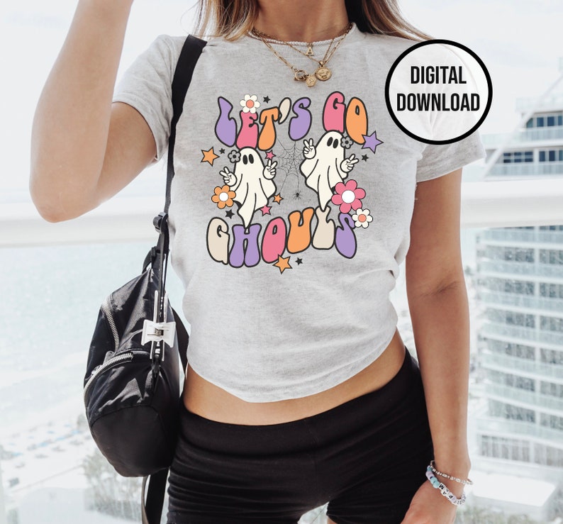 May include: A light grey t-shirt with the text "Let's Go Ghouls" in a retro font, featuring two cartoon ghosts, flowers, and stars. A black circle with "DIGITAL DOWNLOAD" is in the upper right corner. The shirt is part of a Halloween theme.