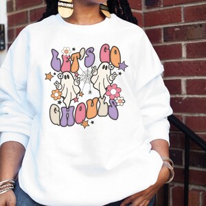 May include: White sweatshirt with a colourful graphic that reads "Let's Go Ghouls." The design includes two cartoon ghosts, flowers, stars, and a spiderweb. The text and images are in shades of orange, purple, pink, and beige.