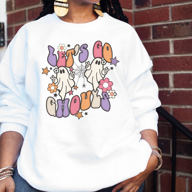 May include: White sweatshirt with a colourful graphic that reads "Let's Go Ghouls." The design includes two cartoon ghosts, flowers, stars, and a spiderweb. The text and images are in shades of orange, purple, pink, and beige.