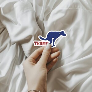 Puede incluir: Una pegatina azul y roja con la silueta de un perro defecando y la palabra "TRUMP" impresa debajo.