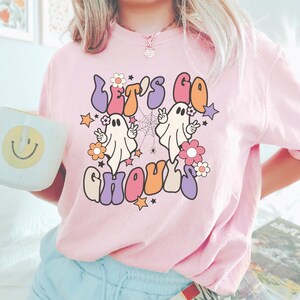 May include: A light pink t-shirt featuring the phrase "LET'S GO GHOULS" in a retro font. The design includes two cartoon ghosts, flowers, stars, and a spider web. The shirt is a casual, comfortable style.