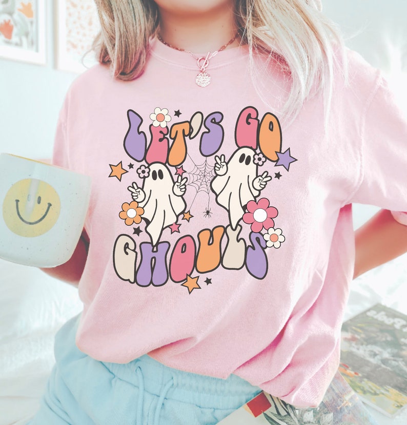 May include: A light pink t-shirt featuring the phrase "LET'S GO GHOULS" in a retro font. The design includes two cartoon ghosts, flowers, stars, and a spider web. The shirt is a casual, comfortable style.