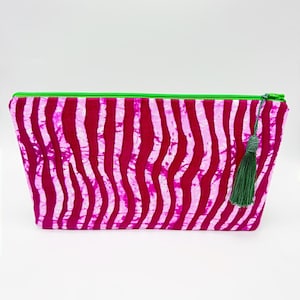 Puede incluir: Un neceser rectangular con un vibrante estampado de rayas verticales onduladas en tonos rosas y blancos. La bolsa presenta una cremallera verde brillante y una borla verde a juego. La bolsa es probablemente de tela.