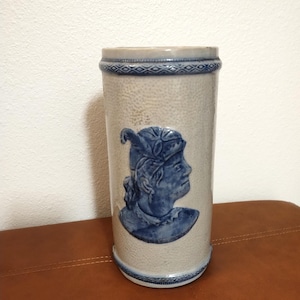 Puede incluir: Un jarro de cerveza cilíndrico de gres, con un fondo beige claro y un perfil azul de una persona. Los bordes superior e inferior tienen bandas decorativas azules. El perfil de la persona también es azul.