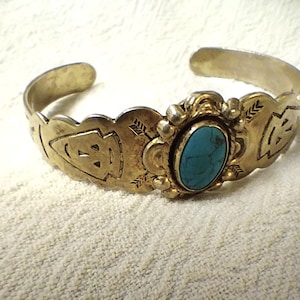 Puede incluir: Un brazalete dorado con una piedra ovalada turquesa en el centro. El brazalete presenta diseños grabados de flechas y geométricos. El brazalete es abierto en los extremos.