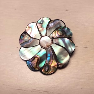 Puede incluir: Broche en forma de flor con pétalos de concha de abulón iridiscente y un centro nacarado. El broche tiene un borde oscuro y refleja tonos verdes, azules y dorados. Un accesorio único para añadir un toque de elegancia inspirado en la naturaleza.